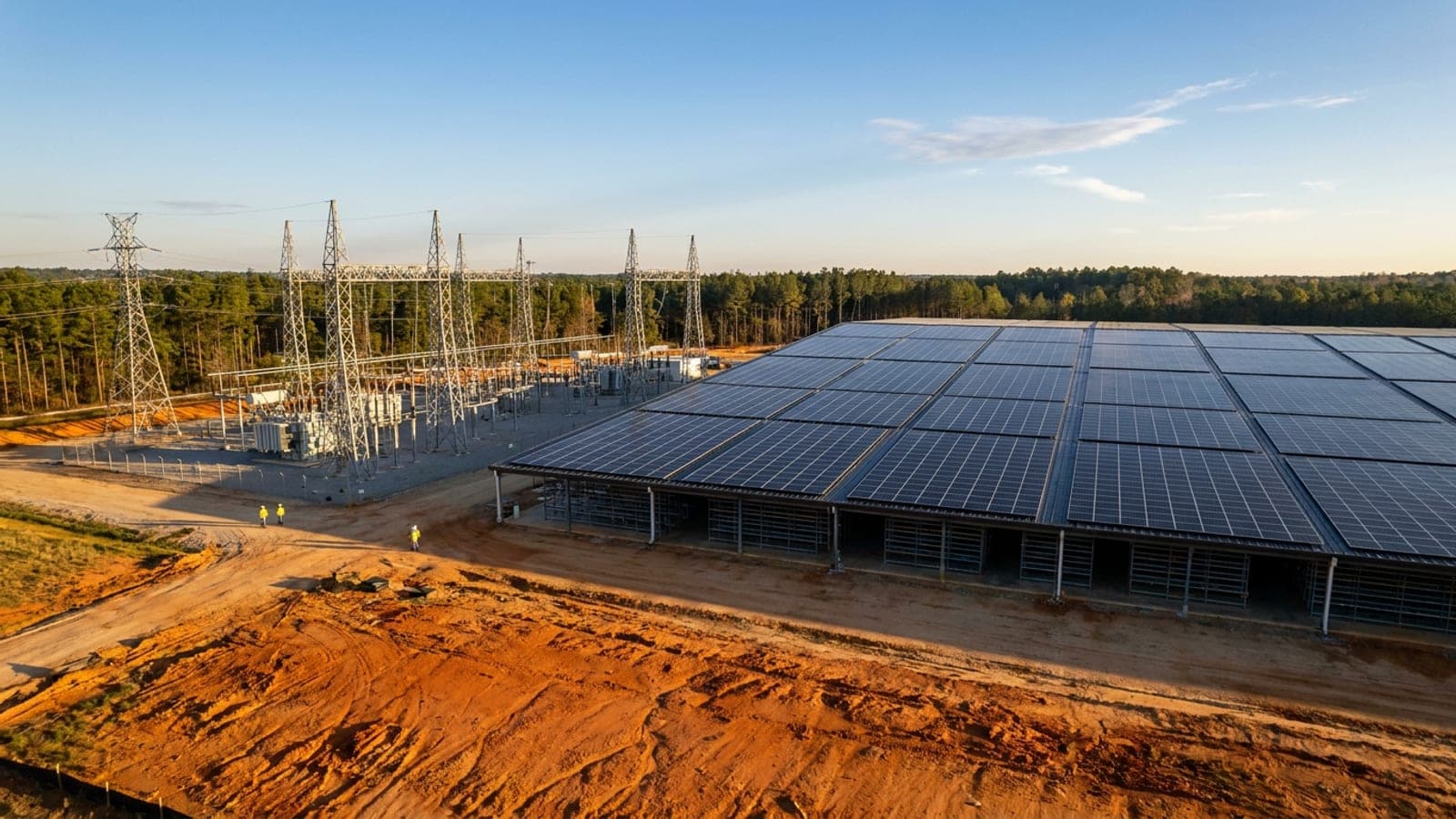 Industrial building with a rooftop solar array beside a utility substation, pine forest behind